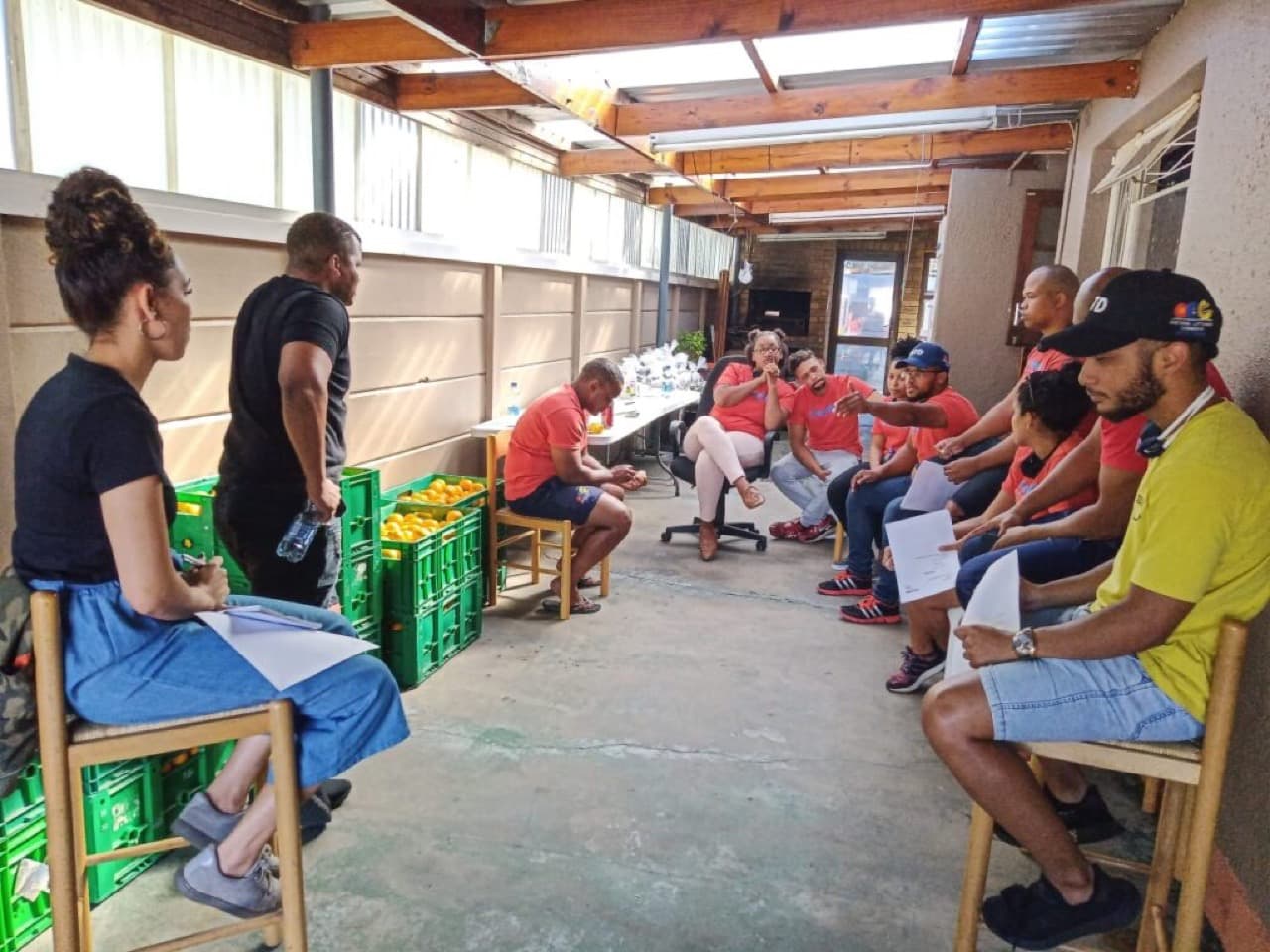 TeamBTD volunteers in red shirts in a planning circle beside crates of oranges for a nutrition programme.