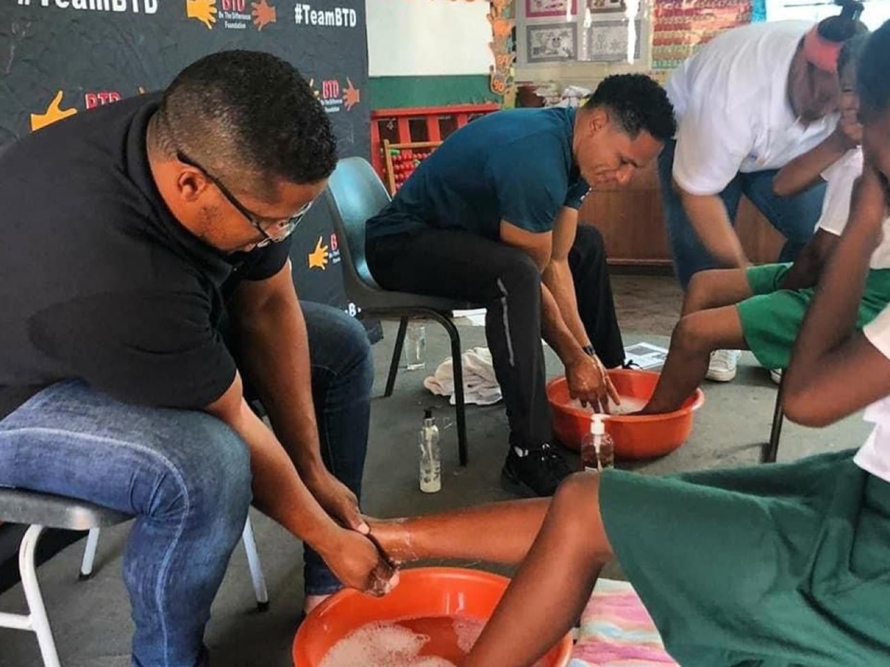 TeamBTD volunteers washing schoolchildren's feet in basins during a school outreach day.