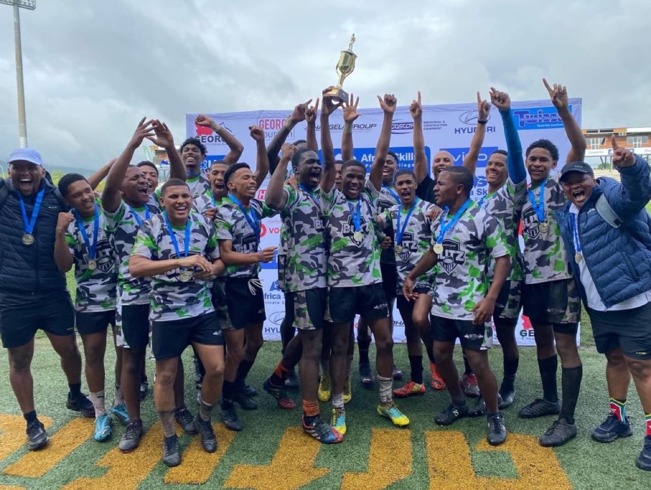 A youth rugby team in BTD-branded camouflage kit celebrating with medals and a trophy.